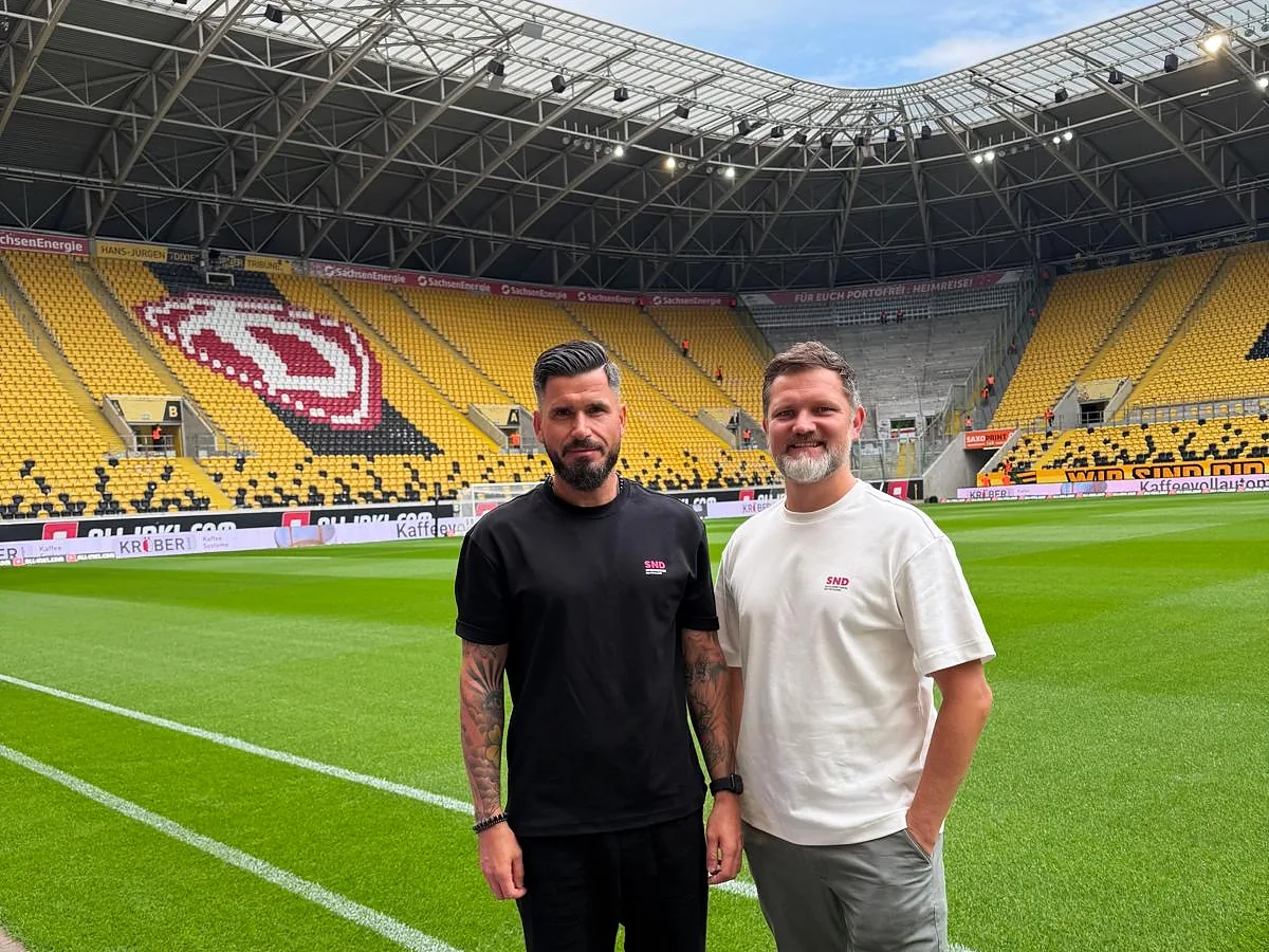 Zwei Personen aus dem SND-Team in SND-Shirts auf dem Rasen des Rudolf-Harbig-Stadions, dahinter gelbe Sitzreihen mit dem Dynamo-Wappen
