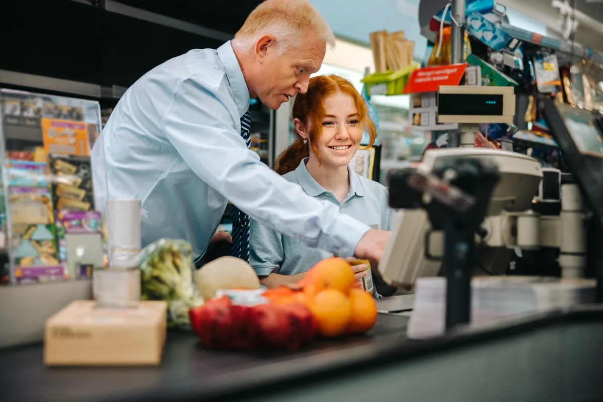 Ein Ausbilder zeigt einer lächelnden Auszubildenden an der Supermarktkasse, wie das Kassensystem funktioniert