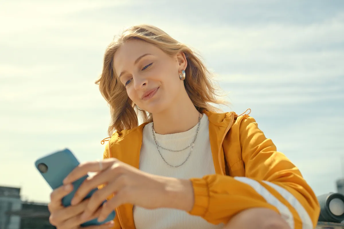 Eine jugendliche Person in gelber Jacke sitzt draußen in der Sonne und schaut lächelnd auf ihr Smartphone