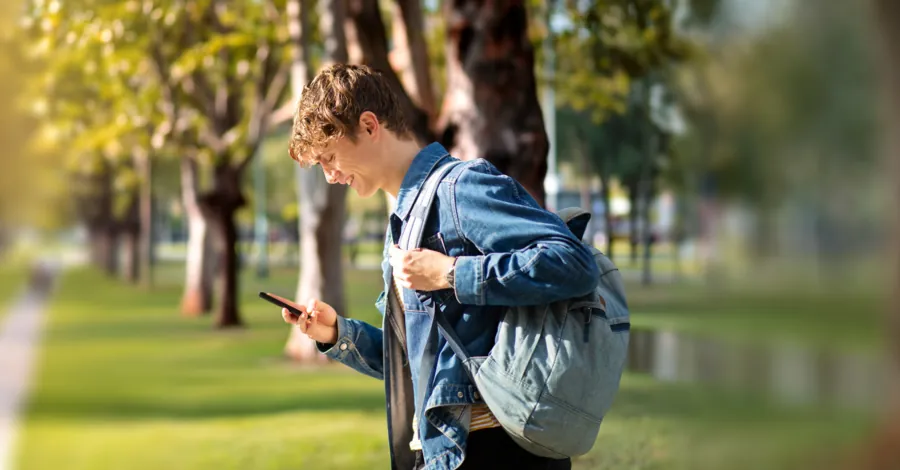 Ein junger Mann mit Rucksack schaut im Park auf sein Smartphone.